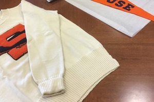 “A white sweatshirt with an embroidered orange letter ‘S’ and an orange textile stating “Syracuse” on top of a brown table.